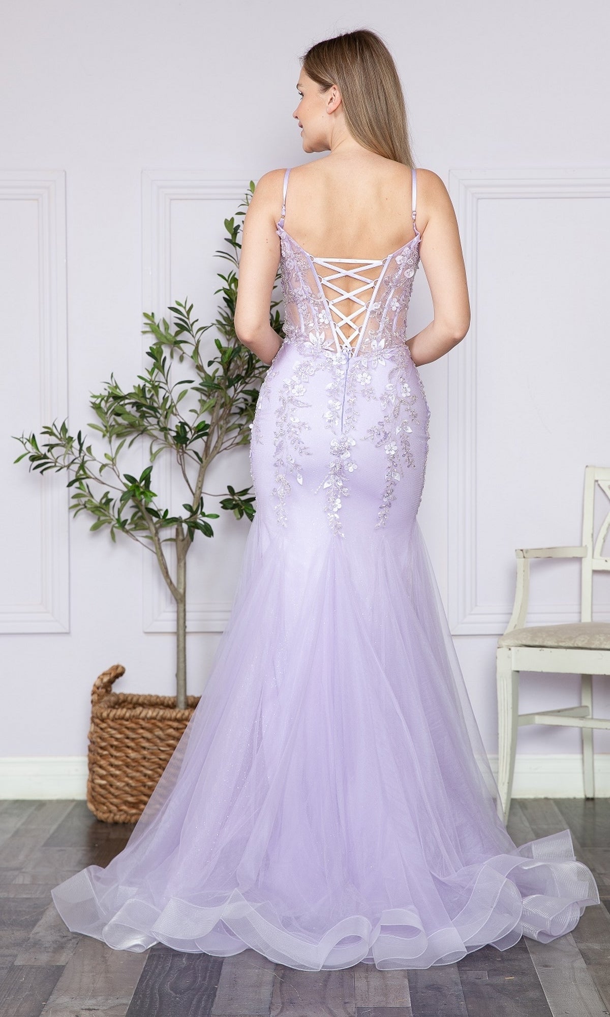 A woman stands indoors near a potted plant and white chair, facing away while wearing the Sheer-Corset-Bodice Long Mermaid Prom Dress 9374—a lilac, floor-length gown with a lace-up back and floral embroidery.